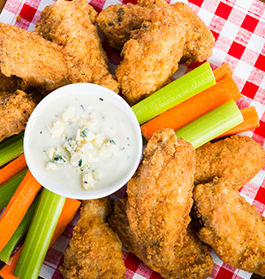 Crispy chicken wings, blue cheese dressing, sliced carrots and celery sitting on top of a red checkered tablecloth