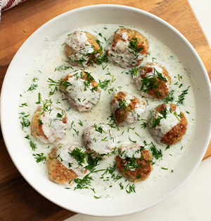 Chicken Meatballs with Yogurt Dill Sauce on a white serving dish sitting on top of a wooden board with a tub of Astro Plain 6% yogurt beside it.